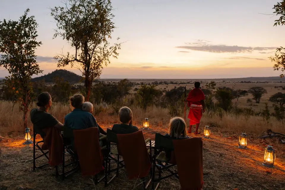 Guests seated around lanterns in the bush at sunset with a Maasai guide