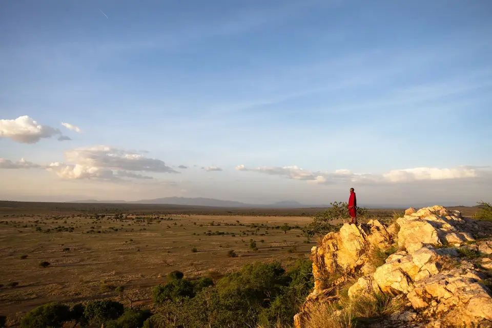 A Maasai guide in a red shuka stands on a rock outcrop above the golden plains of Randilen at dusk